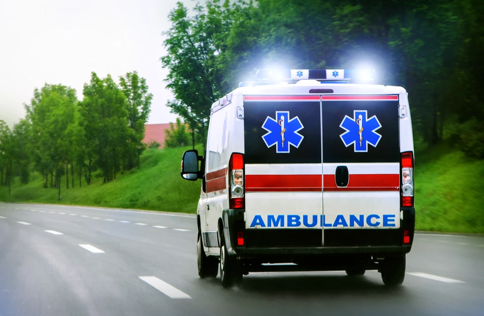 Ambulance en intervention circulant sur route avec gyrophares activés, véhicule sanitaire blanc aux insignes médicaux bleus, bandes réfléchissantes rouges, en environnement rural verdoyant