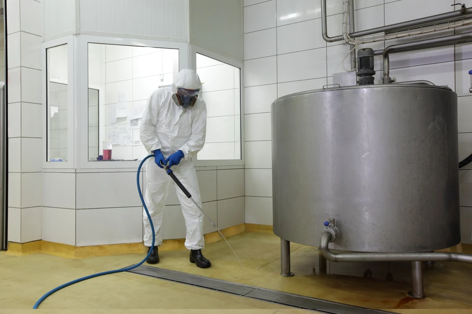 Professionnel en combinaison de protection intégrale grise avec masque respiratoire effectue une désinfection par pulvérisation dans un hall d'immeuble près d'un ascenseur