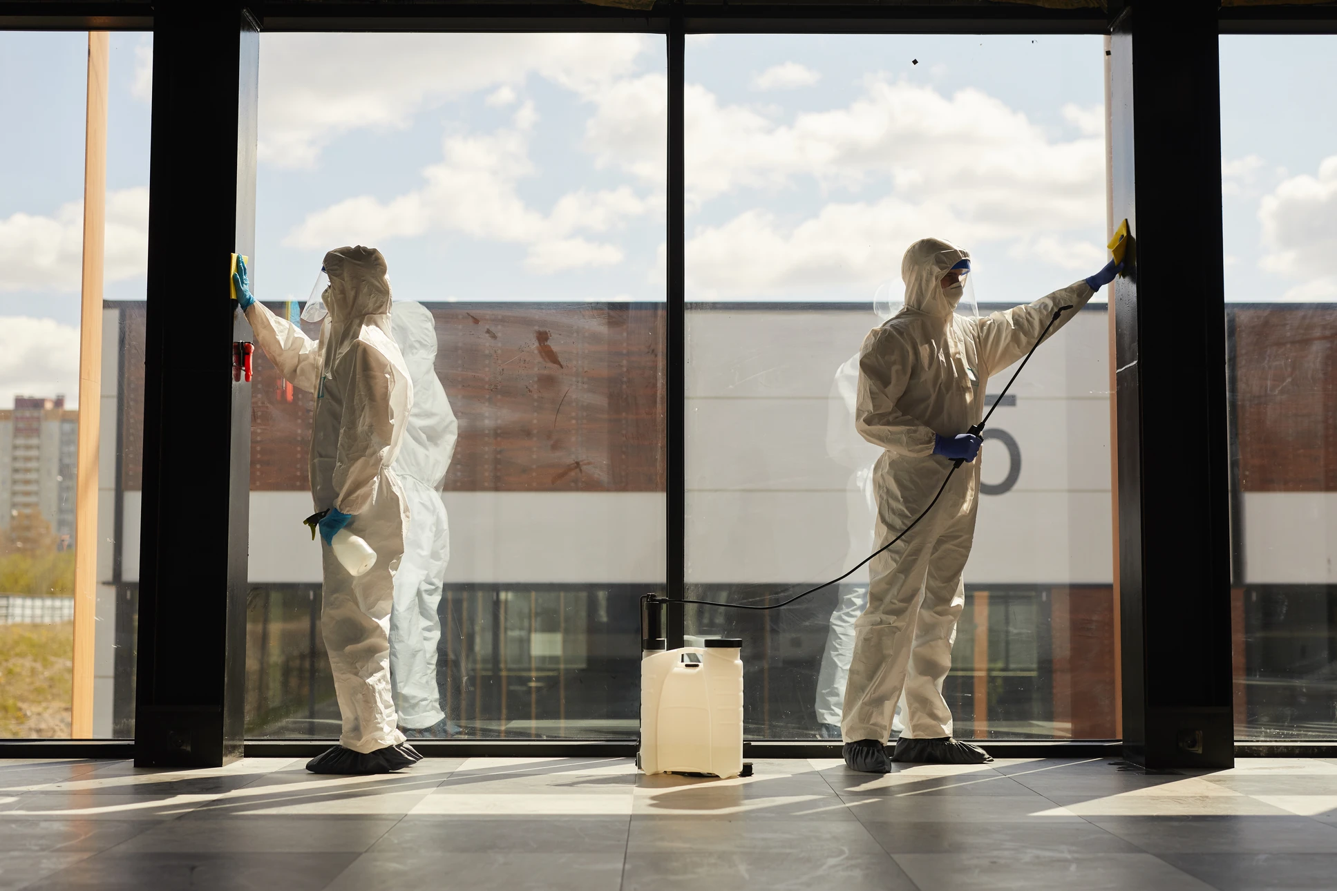 Professionnel en combinaison de protection intégrale grise avec masque respiratoire pulvérise un désinfectant dans un couloir d'immeuble près d'un ascenseur